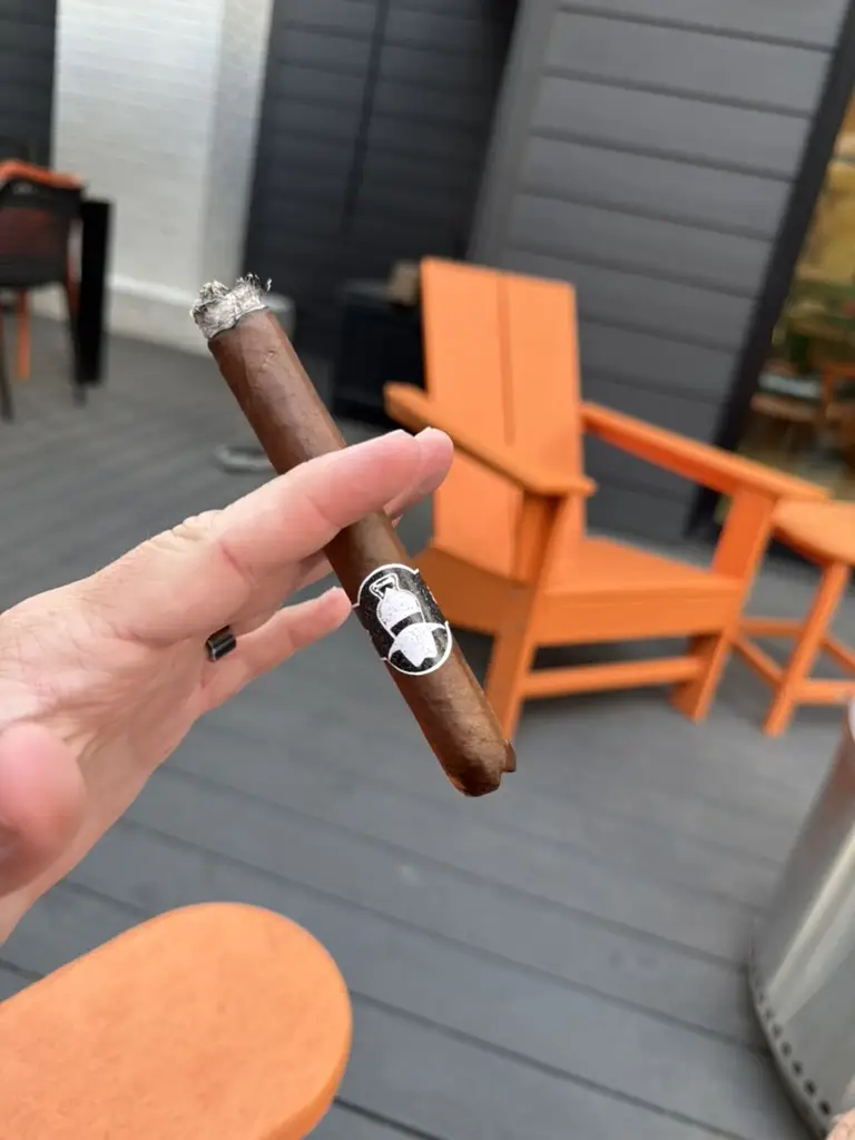 A three year old SLAB Cigars stick with a cartoon band, burning clean with a tight grey ash, held over the deck against an orange Adirondack chair