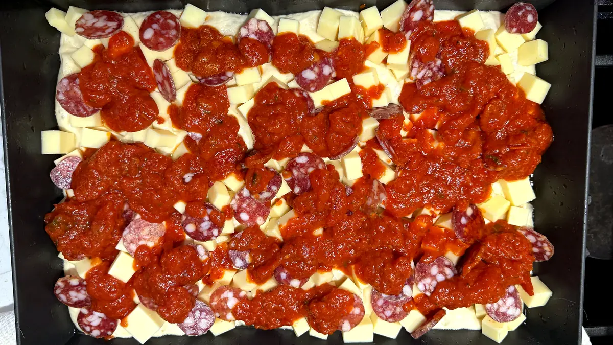 Detroit pizza assembled in the pan before baking, cubed cheese pushed to the edges with flat salami coins and dolloped sauce on top
