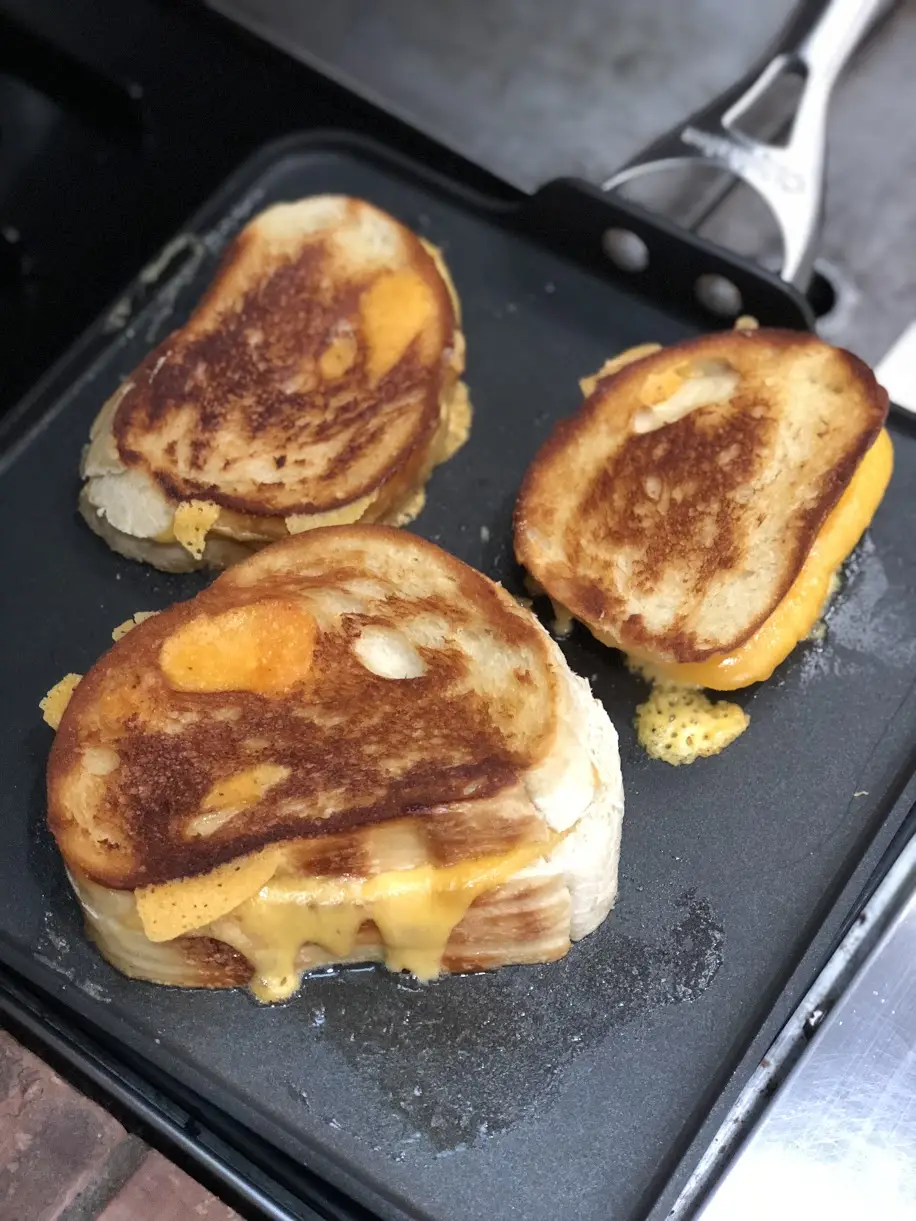 Three grilled cheese sandwiches on a black griddle with cheese oozing out and crisping on the surface