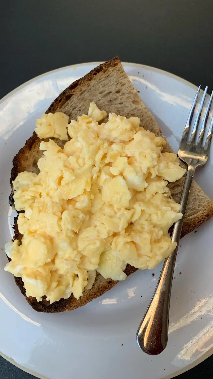 Soft scrambled eggs in loose folds over a thick slice of dark toasted sourdough on a white plate