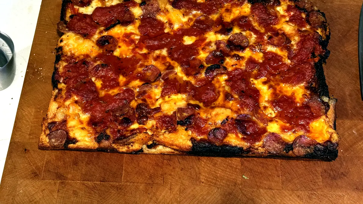 Baked Detroit-style pizza on a wooden cutting board, dark crispy charred cheese edges, sauce and salami on top